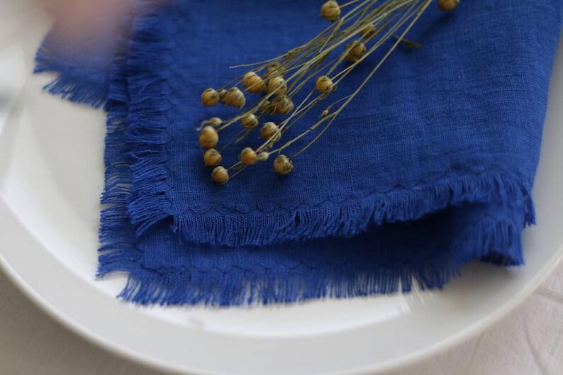 May include: A blue linen napkin with frayed edges, folded and placed on a white plate. A sprig of dried flax flowers with seed pods is placed on top of the napkin.