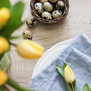 May include: A blue linen napkin with a frayed edge is folded on a white plate. A single yellow tulip with green leaves is placed on top of the napkin. A nest of quail eggs is in the background.