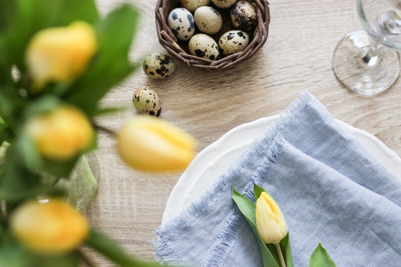 May include: A blue linen napkin with a frayed edge is folded on a white plate. A single yellow tulip with green leaves is placed on top of the napkin. A nest of quail eggs is in the background.