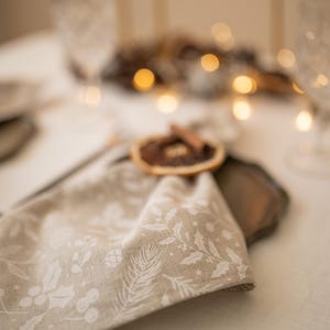 May include: A beige linen napkin with a white floral pattern is tied with a cinnamon stick and dried orange slice. The napkin is on a table setting with a brown plate and a white tablecloth.