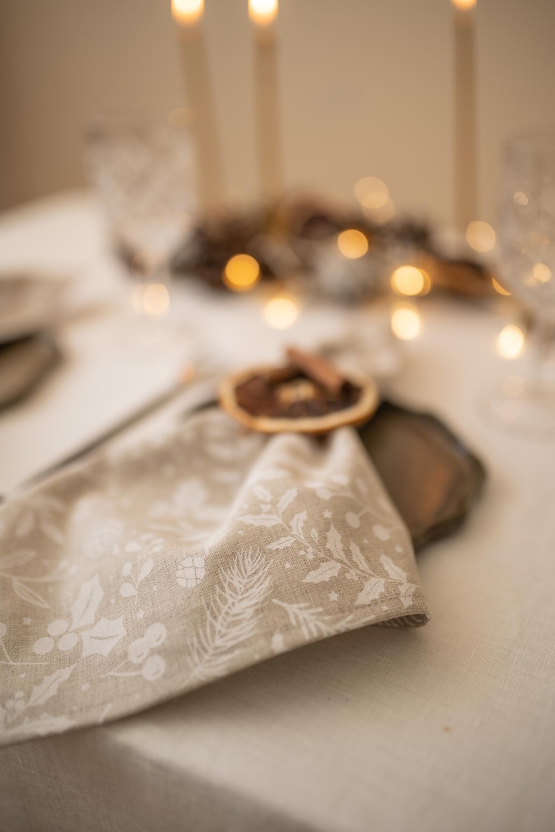 May include: A beige linen napkin with a white floral pattern is tied with a cinnamon stick and dried orange slice. The napkin is on a table setting with a brown plate and a white tablecloth.