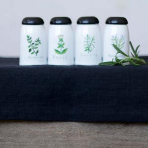 Op de afbeelding: Vijf witte keramische kruidenpotten met zwarte deksels en groene kruidenillustraties. De potten zijn in een rij op een zwarte linnen loper geplaatst.