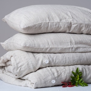 Può includere: Un mucchio di quattro federe di cuscino in lino a righe di colore beige chiaro e marrone. Le federe sono piegate e disposte su uno sfondo grigio.
