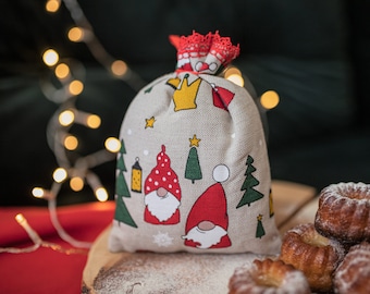 Puede incluir: Una bolsa de regalo de tela beige con un borde de encaje rojo y blanco. La bolsa está decorada con un tema navideño, con gnomos, estrellas y árboles. La bolsa está sentada sobre una superficie de madera con azúcar en polvo.
