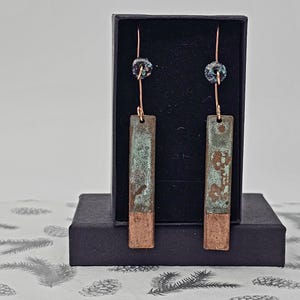 May include: A pair of long, rectangular earrings with a copper and blue patina finish. The earrings are hanging from a black box.