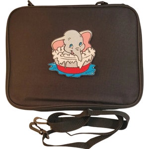 以下が含まれることがあります： 黒色の長方形のキャリングケース。ファスナー開閉式で、取り外し可能な黒色のショルダーストラップが付いています。ケースには、赤い浴槽に入ったダンボの刺繍パッチがあり、青い水に囲まれています。