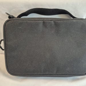 May include: A rectangular black bag with a black handle and a zipper closure. The bag has a textured surface and a small loop on each side. The bag is designed to carry items.