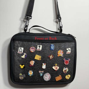 May include: Black rectangular bag with a shoulder strap, covered in colorful enamel pins. The pins feature various cartoon characters and designs. The text "Front or Back" is printed in red on the bag.