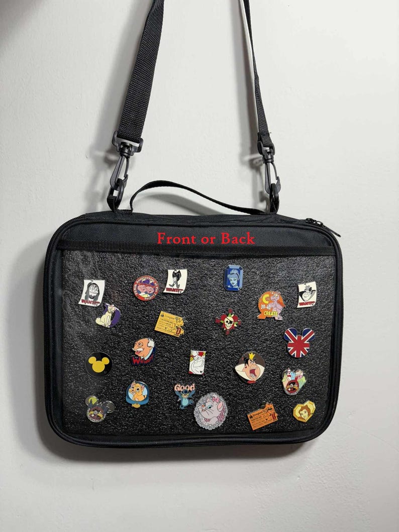 Puede incluir: Bolso rectangular negro con una correa para el hombro, cubierto de varios pines esmaltados. Los pines presentan personajes de dibujos animados y otros dise&ntilde;os. El texto "Front or Back" est&aacute; impreso en rojo en el bolso.