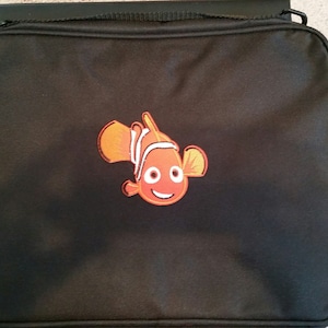 May include: Black rectangular bag with a zipper closure and a small orange and white embroidered fish design on the front. The bag has a handle on top.