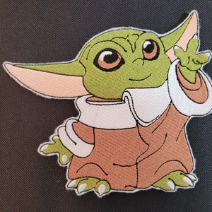 Puede incluir: Parche bordado con un personaje alien&iacute;gena verde con orejas grandes, ojos marrones y una t&uacute;nica marr&oacute;n. El personaje sonr&iacute;e y tiene una mano levantada. El parche tiene un borde blanco y est&aacute; sobre un fondo gris oscuro.