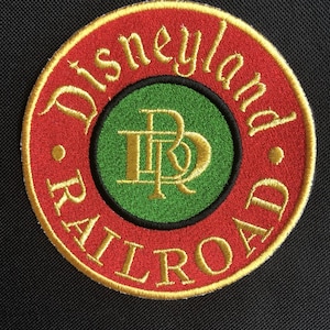 May include: A red and gold embroidered patch with the text "Disneyland Railroad" in a circle. The center of the patch features a green circle with a gold "D" and "R" intertwined.