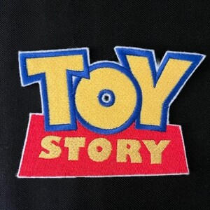 May include: Embroidered patch featuring the "Toy Story" logo. The text "TOY STORY" is in yellow with blue outlines, set on a red rectangular base. The patch is against a black background.