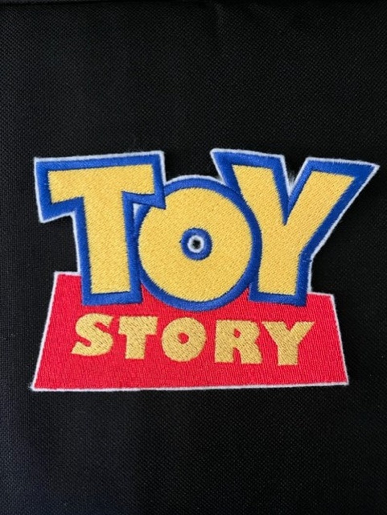 May include: Embroidered patch featuring the "Toy Story" logo. The text "TOY STORY" is in yellow with blue outlines, set on a red rectangular base. The patch is against a black background.
