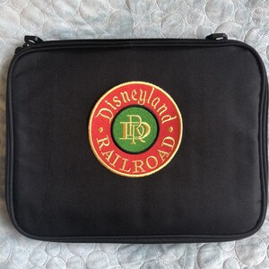 May include: Black rectangular case with a Disneyland Railroad logo. The logo is a circular patch with red, green, and gold lettering and design. The case has a zipper closure and two small black handles.