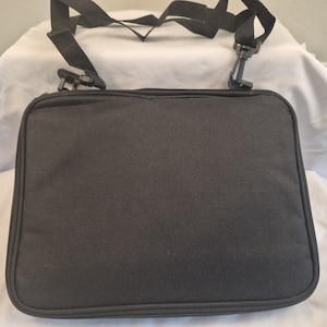 May include: A black rectangular bag with a shoulder strap. The bag has a zipper closure and a textured surface. The adjustable strap is attached with black plastic clips.