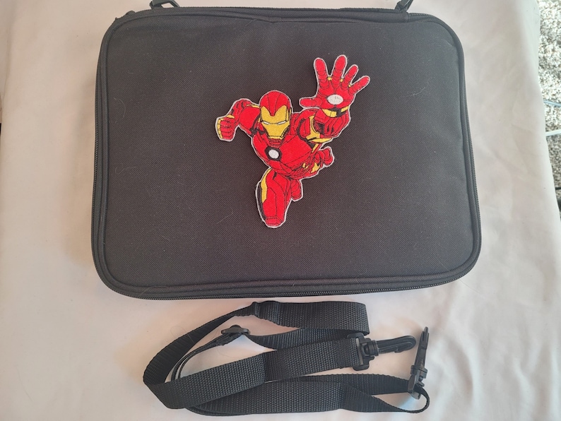 May include: Black rectangular bag with a red and yellow Iron Man patch. The bag has a zipper closure and a detachable black shoulder strap. The Iron Man patch depicts the superhero in a dynamic pose.