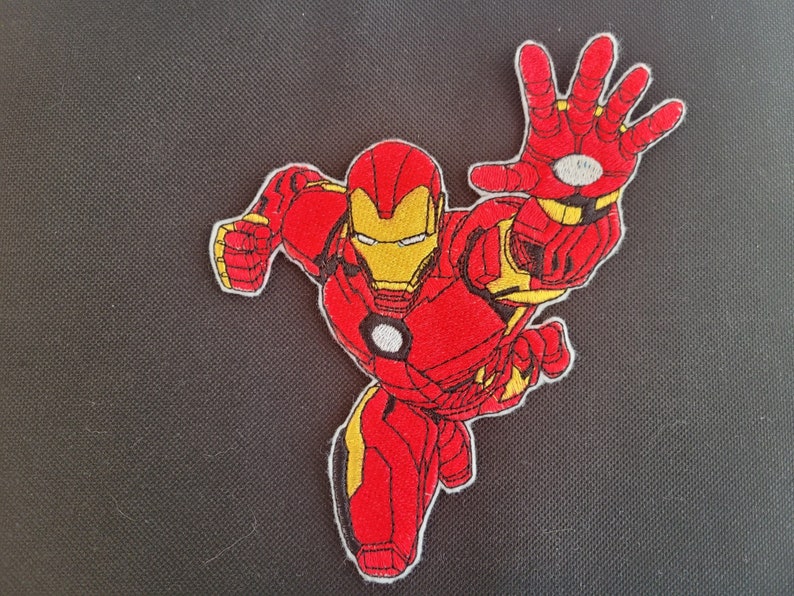 May include: Embroidered patch featuring Iron Man in a dynamic pose, with his right hand extended. The patch showcases a red and yellow color scheme, with detailed stitching outlining the superhero's iconic suit. The patch is likely for clothing or accessories.
