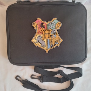 May include: A black rectangular zippered bag featuring a central embroidered crest with a large letter H surrounded by a lion, a snake, a badger, and an eagle. The bag includes a detachable black adjustable shoulder strap with plastic clips.