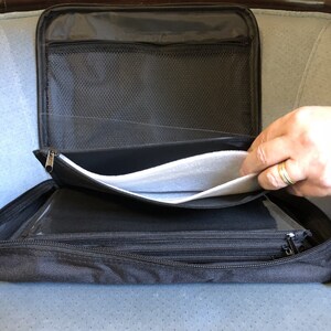 May include: Black zippered organizer case with multiple compartments. The case is open, revealing a mesh pocket, clear plastic sleeves, and a padded interior. A hand with a ring is visible.