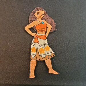 Puede incluir: Parche bordado que representa un personaje de dibujos animados con vestimenta tradicional. El personaje tiene cabello casta&ntilde;o, un top naranja y una falda blanca con dise&ntilde;os de flores naranjas. Se ve un collar de gemas azules. El parche est&aacute; sobre un fondo negro.