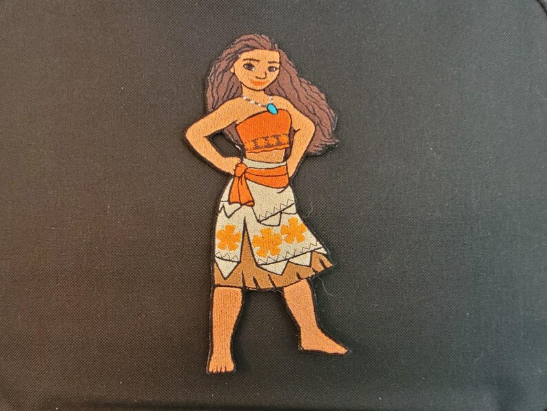 Puede incluir: Parche bordado que representa un personaje de dibujos animados con vestimenta tradicional. El personaje tiene cabello casta&ntilde;o, un top naranja y una falda blanca con dise&ntilde;os de flores naranjas. Se ve un collar de gemas azules. El parche est&aacute; sobre un fondo negro.