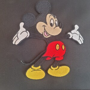 May include: Black bag featuring an embroidered image of Mickey Mouse. The cartoon character is depicted with a black head and ears, a tan face, white gloves, red shorts, and yellow shoes. The bag is likely made of a durable fabric.
