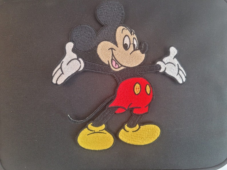 May include: Black bag featuring an embroidered image of Mickey Mouse. The cartoon character is depicted with a black head and ears, a tan face, white gloves, red shorts, and yellow shoes. The bag is likely made of a durable fabric.