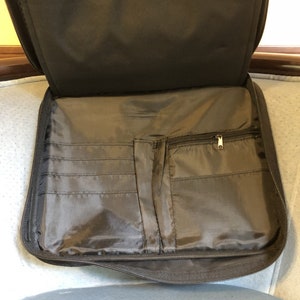 May include: Open, dark gray zippered portfolio with multiple interior pockets and a zippered compartment. The portfolio is made of a smooth, durable material and is designed to hold documents and other items. The interior features several card slots.