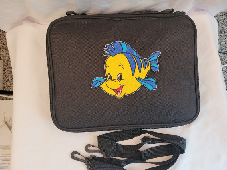 May include: Black rectangular case with a zipper closure and a Flounder patch. The patch features a yellow fish with blue accents. Includes a black shoulder strap with two clips.