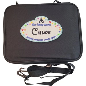 May include: Black travel bag with a detachable shoulder strap. The bag features a patch with the text "Chloe" and "Where Dreams Come True" with a castle and star design. The bag has a zipper closure.