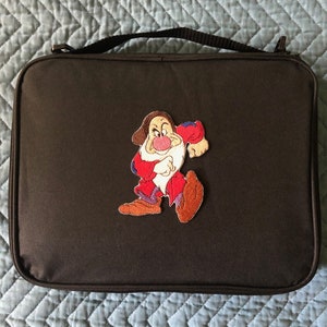 May include: A black rectangular case with a handle and a zipper closure. The case features an embroidered patch of a cartoon character in red, white, and brown clothing. The character has a large nose and a grumpy expression.