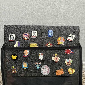 May include: A collection of enamel pins, featuring various cartoon characters and designs. The pins are attached to a black display case with a clear front. Some pins have text, including "WANTED" and "Good".