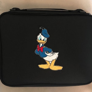 May include: Black travel case featuring an embroidered image of Donald Duck. The cartoon character is depicted in his signature blue sailor shirt, bow tie, and hat. The case has a handle and a zipper closure.