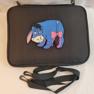 May include: Black rectangular bag featuring a cartoon image of Eeyore from Winnie the Pooh. The blue donkey has a pink bow and purple tail. A black shoulder strap with clips is included.
