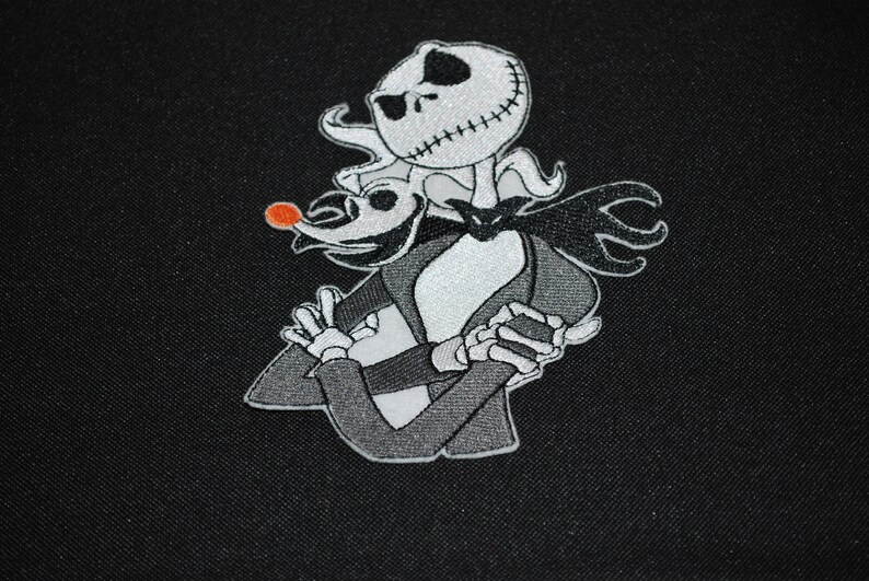 Puede incluir: Parche bordado con Jack Skellington y Zero de Pesadilla antes de Navidad. Jack, con una cara cosida, lleva un traje negro, Zero tiene una nariz roja. El parche es en tonos negros, blancos y grises.