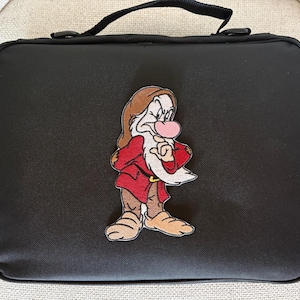 May include: A rectangular black lunch bag with a top handle and a zipper closure. Centered on the front is an embroidered patch of the character Grumpy from Snow White, featuring a red tunic, brown hat, white beard, and a pink nose.