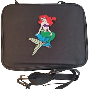 May include: Black rectangular case with a zipper closure and a detachable shoulder strap. The case features an embroidered image of a mermaid with red hair, a purple top, and a green tail. The mermaid is sitting on a blue flower.