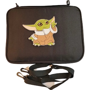 Puede incluir: Bolso rectangular negro con una imagen bordada de un personaje alien&iacute;gena verde con ojos grandes y orejas puntiagudas. El bolso tiene una correa negra para el hombro con dos clips negros.