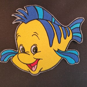 May include: Embroidered patch featuring a cartoon fish with a yellow body, blue fins, and a smiling red mouth. The fish has large green eyes and is outlined in white. The patch is likely for clothing or accessories.
