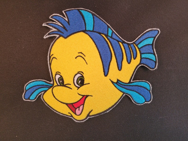 May include: Embroidered patch featuring a cartoon fish with a yellow body, blue fins, and a smiling red mouth. The fish has large green eyes and is outlined in white. The patch is likely for clothing or accessories.