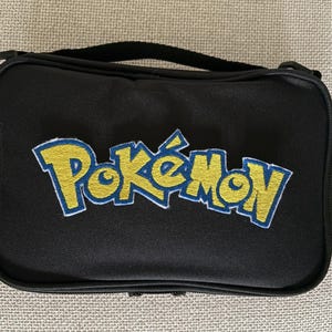 May include: A rectangular black zippered pouch featuring the yellow and blue embroidered Pokémon logo centered on the front. The bag includes a black fabric handle attached to the top edge and a small side loop for carrying or attachment purposes.