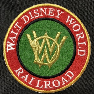Pode incluir: Um remendo bordado em vermelho, dourado e verde com o texto "Walt Disney World Railroad" em branco. O centro do remendo apresenta um "W" estilizado em dourado.