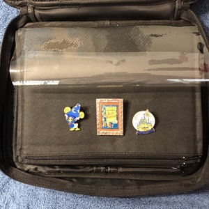 May include: A black zippered carrying case, open to reveal clear plastic sleeves. Three enamel pins are displayed: Donald Duck as a sorcerer, a "Silly Symphonies" pin, and a pin with a castle and "Disney" text. The case is on a blue surface.