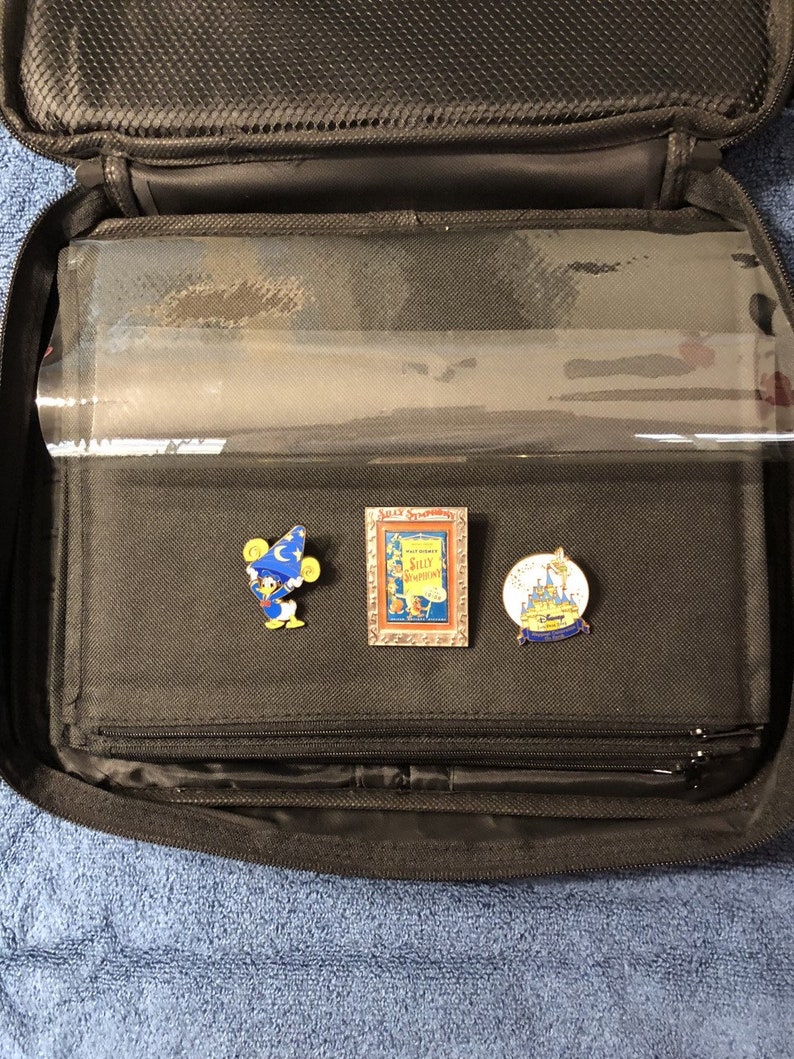 May include: A black zippered carrying case, open to reveal clear plastic sleeves. Three enamel pins are displayed: Donald Duck as a sorcerer, a "Silly Symphonies" pin, and a pin with a castle and "Disney" text. The case is on a blue surface.