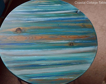 Pode incluir: Uma mesa redonda de madeira pintada em tons de azul, verde e branco, criando um padrão listrado. A mesa tem um estilo rústico e de casa de campo costeira.