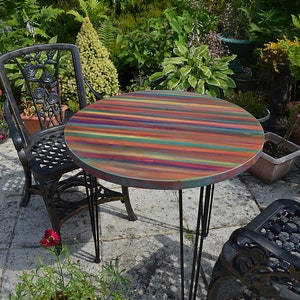 May include: A round, multi-coloured wooden table with black hairpin legs. The table is set on a patio with a black metal chair and a potted plant with red flowers.