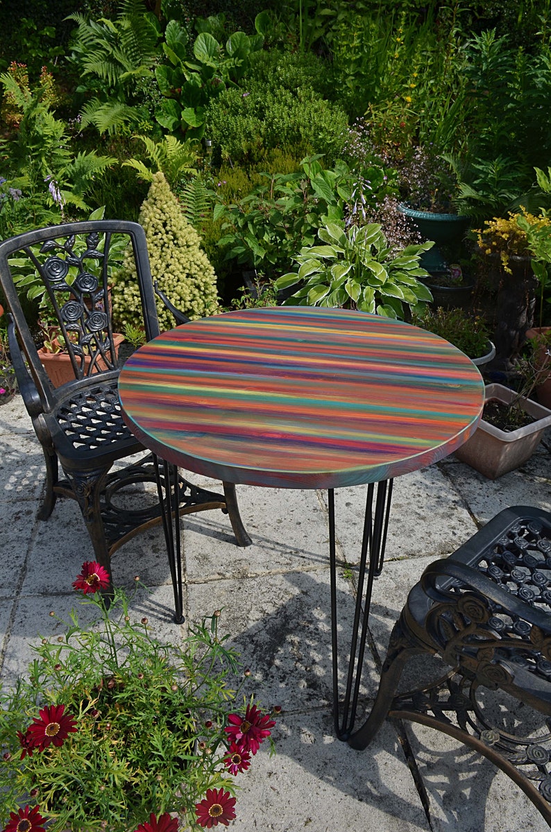 May include: A round, multi-coloured wooden table with black hairpin legs. The table is set on a patio with a black metal chair and a potted plant with red flowers.