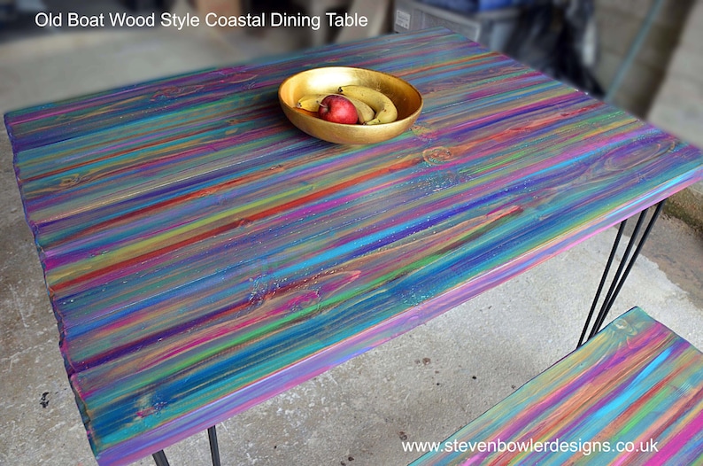 May include: A colourful wooden dining table with a rainbow-like finish. The table has a rustic, coastal style and is made of reclaimed wood. The table is supported by black metal hairpin legs.
