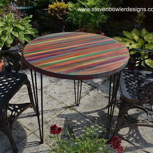May include: A round wooden table with a colourful striped top and black hairpin legs. The table is set up outdoors with two black metal chairs.